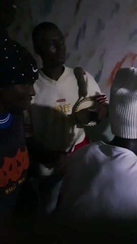 The video appears to capture a gathering or celebration in Barakin Angundi, Nigeria. The scene is dimly lit, suggesting it might be evening or indoors. The subjects are primarily people, with at least three individuals clearly visible. One person in the foreground is wearing a light-colored long-sleeved shirt with red stripes on the sleeves. This individual is holding an object near their face, possibly a microphone or a decorative item. Another person is lying down in the lower part of the frame, wearing a patterned headwrap. A third person, partially obscured, is standing behind the seated individual.

The background shows a textured wall with abstract, colorful markings. There is a sense of social interaction, possibly a performance or a communal event. The emotions conveyed are difficult to ascertain due to the low light and the angle of the recording, but the presence of multiple people suggests a social gathering.

Visible text includes "OFF" and possibly "GUESS" in a gold-colored font on the shirt of the person in the foreground. There are also other indistinct markings on the wall that might be graffiti or decorations. The overall impression is of a lively, informal gathering in a community setting.
