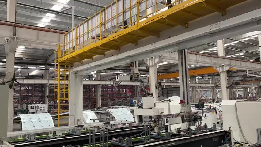 The video showcases a large, brightly lit factory or industrial facility. A complex automated machinery system, primarily white with robotic arms, is the central focus. It operates along a conveyor belt, processing what appear to be foil packages patterned with repeating images. The factory’s framework includes structural columns and a yellow elevated walkway, accessible by a ladder. In the background, partially obscured by the machinery, are stacked shelves and industrial components, suggesting warehousing or inventory storage. The scene includes workers who are minimally visible near the machinery. The overall impression is one of a technologically advanced, automated production environment.