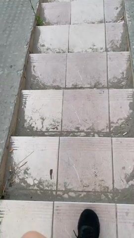 A first-person perspective video shows a person walking down a set of outdoor stairs. The stairs are tiled with light-colored squares, and the edges of the steps are lined with sand or dirt. Numerous cigarette butts are scattered on the steps, particularly along the edges. The person is wearing black sneakers and is partially visible from the waist down, with their right leg stepping forward. To the left of the stairs, there is a grassy area with a single green plant. The edges of the stairs appear to be made of a textured gray material. The video pans down the stairs as the person descends.