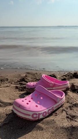 The video shows a pair of pink Croc shoes resting on a sandy beach with the ocean in the background. The shoes are positioned close together on the sand, with the left shoe slightly behind the right. The ocean water is calm with small waves lapping at the shore. The sky is a clear blue with a few white clouds. The sun appears to be shining, casting shadows on the sand. The overall scene suggests a relaxed, summery atmosphere at the beach.