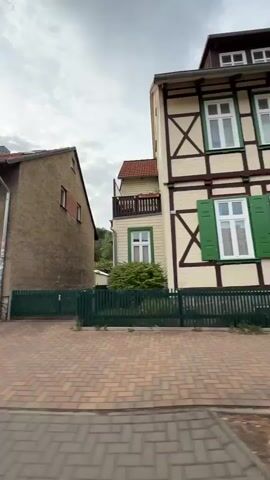 The video pans across a street with buildings and a sidewalk. The first building is a light brown house with white-trimmed windows and a small wooden fence. A rose bush with pink flowers is visible in front of it. Next, a large wooden gate is shown, situated between two buildings, one with a half-timbered facade and the other with a stucco exterior. Finally, the camera moves to show a row of buildings with shops on the ground floor. One shop has a sign that reads "Coiffeur". A man in a light-colored t-shirt and shorts walks by the shop. The sky is overcast.