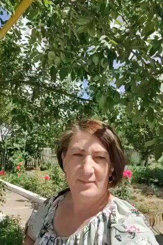 A woman with short brown hair is standing in front of a tree. She is wearing a light-colored shirt with a floral pattern. The background is a garden with pink roses. The sky is blue with some clouds. The video is shaky and appears to be a selfie.