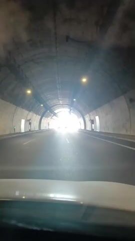The video shows a POV shot from inside a car driving through a tunnel. The tunnel is made of concrete and is lit by overhead lights. The car is moving towards a bright light at the end of the tunnel, which indicates the exit. The road inside the tunnel is asphalt with white lane markings. There are some structures on the sides of the tunnel, and lights mounted on the ceiling. As the car gets closer to the exit, the view opens up to reveal daylight and other cars on the road ahead. The video is short and seems to capture the transition from the darkness of the tunnel to the brightness of the outside.