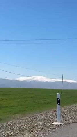 A video taken from inside a moving vehicle shows a snow-capped mountain in the distance against a clear blue sky. In the foreground, there is a green field and a grey metal guardrail. A few trees, some bare and some with sparse leaves, line the roadside. The camera is shaky due to the movement of the vehicle. Power lines can be seen stretching across the field towards the mountains. Road signs appear fleetingly.