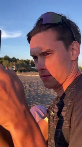 A close-up shot shows a young man's face illuminated by the warm glow of the sun. He is wearing sunglasses pushed up on his forehead and a patterned t-shirt. His gaze is directed downwards, focused on something out of frame, possibly a phone he is holding. His left arm is partially visible, also catching the sunlight. In the background, a beach is visible with the sky overhead and distant foliage. Another person, wearing a light-colored t-shirt with green lettering, is visible in the background behind the main subject. The overall impression is a candid moment captured outdoors, possibly during sunset.