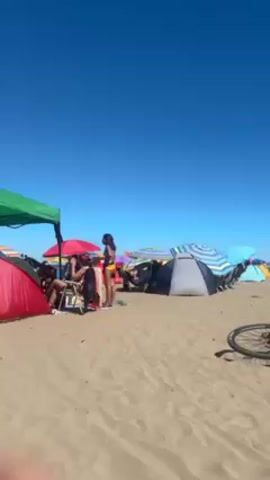 The video shows a sunny beach scene with people relaxing and socializing. There are several colorful beach umbrellas and tents dotting the sandy landscape. People are sitting in chairs, standing around, and walking across the beach. Some individuals are wearing swimwear. The sky is clear and blue.