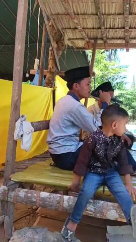 A young boy in a patterned shirt and blue jeans sits on a wooden structure, facing right, with his legs dangling. Behind him, a young man wearing a black peci and a striped shirt holds a microphone and appears to be speaking. He is seated on a mat with a green and white floral pattern. To his left, another person, partially obscured, is also seated. In the background, several other individuals are visible, including a man in a brown shirt seated further back. The scene is outdoors, under a thatched roof supported by wooden beams. Yellow fabric is visible behind the seated figures. The overall environment suggests a casual outdoor gathering or event.