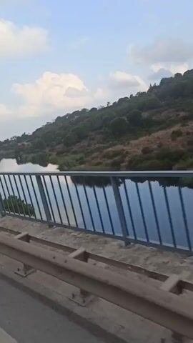 The video shows a view from a bridge looking out over a body of water. A green, tree-covered hill rises from the far bank of the water. The sky is blue with scattered white clouds. The camera is moving, indicating the view is from a vehicle on the bridge. The bridge has a metal railing and a guardrail.