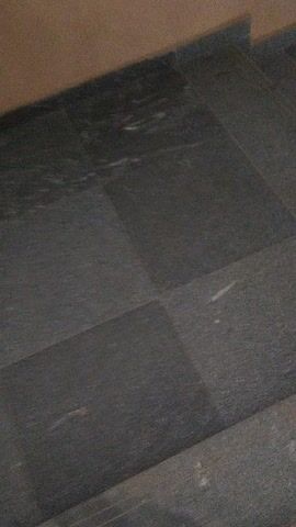 The video opens with a very close, blurry shot of a dark grey or black textured surface, possibly a floor or wall, with faint lighter streaks. On the left side of the frame, a blurry, light brown or beige object, which appears to be a human limb (likely a leg or arm), is visible. The camera then begins to pan downwards and slightly to the right. As it moves, a series of dark grey steps with a rough, stone-like texture become visible, indicating a staircase. The blurry light brown object continues to move along the left edge of the frame, suggesting a person is walking down the stairs while being filmed from a close perspective. The lighting in the scene appears to be somewhat dim. The video ends with a blurry view of the steps and the moving limb.