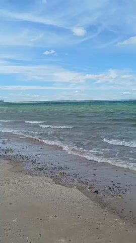 A video clip shows a wide shot of a beach in Redcar, United Kingdom. The scene is dominated by the ocean, with waves gently lapping at the shore. The water is a greenish-blue, and the sky above is a clear blue with scattered white clouds. The beach itself is composed of sand and pebbles, with some seaweed visible along the waterline. In the distance, there are a few buoys or markers in the water, and what appears to be a wind turbine on the horizon. The overall mood is peaceful and serene, suggesting a pleasant day at the seaside. The time of day appears to be midday, given the bright sunlight and clear skies. There is no text visible in the video.
