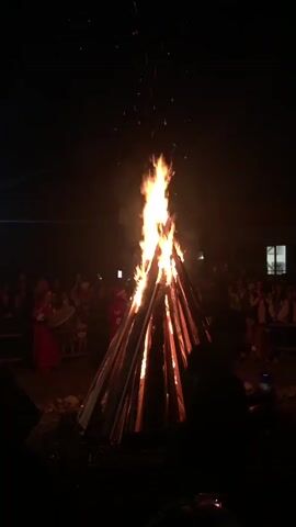 A large bonfire is lit at night. The bonfire is cone-shaped and made of logs. Sparks fly from the top of the fire. A crowd of people are gathered around the bonfire. Some of the people are wearing traditional clothing. The background is dark and indistinct.