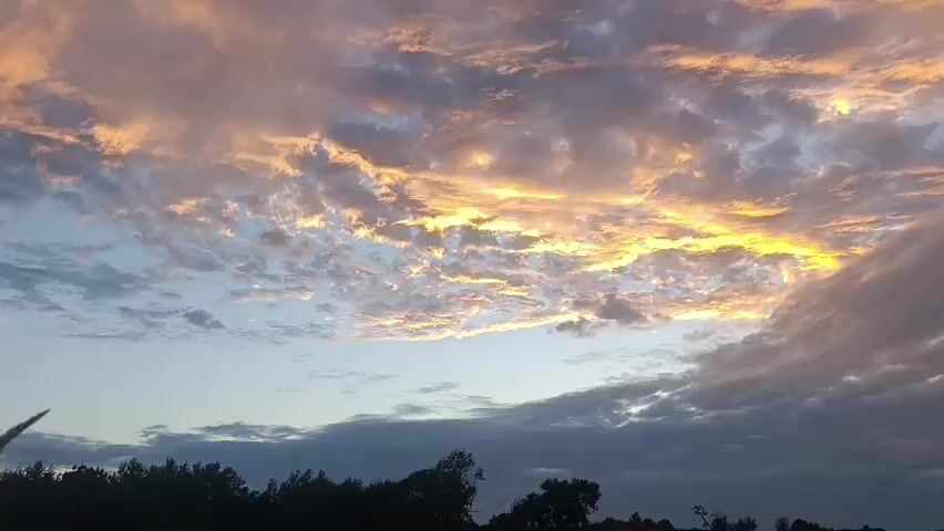 A wide shot captures a sunset sky filled with clouds. The clouds are illuminated by the setting sun, with streaks of orange and yellow light visible in the upper portion of the frame. The lower portion of the sky is a darker blue, and a silhouette of trees and a hilly landscape is visible along the bottom edge of the image. The clouds appear to be moving slowly across the sky.
