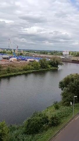 The video shows a wide river with a dark surface. On the left bank of the river, there is an industrial area with several buildings, a large crane, a stack of blue cylindrical objects, and some vehicles. Further inland, two wind turbines are visible. On the right bank of the river, there is a concrete structure that appears to be part of a dam or lock system. The sky is overcast with clouds. The foreground shows green foliage and a road. The camera is positioned at an elevated angle, looking across the river.