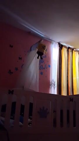 A Spongebob Squarepants toy hangs from a string above a white crib. The crib has a decorative crown on the side. The wall behind the crib is pink and decorated with blue stars and butterflies. Curtains are visible in the background.