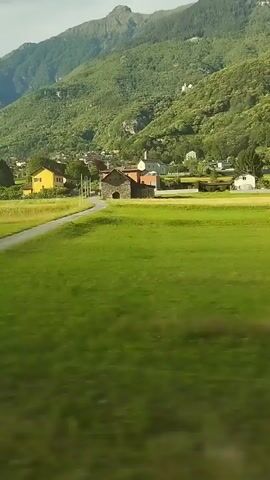The video shows a landscape with green fields and mountains in the background. There are some buildings visible in the distance, and trees are scattered throughout the scene. The camera appears to be moving, possibly from a vehicle, as evidenced by the blur effect in some parts of the video, particularly the foreground. The lighting suggests it is daytime, possibly late afternoon given the angle of the sun. The overall impression is of a rural, mountainous region.