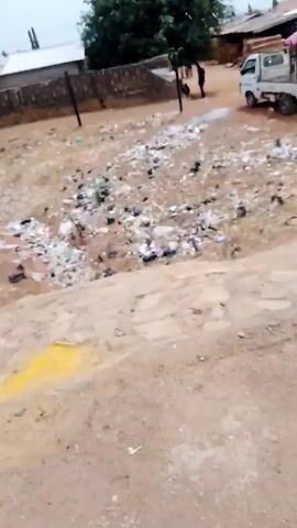The video shows a sandy, unpaved ground covered in trash, mostly black plastic bags and white plastic debris. A small, green bush is present amidst the litter. In the background, there is a low brick wall with metal poles running along its top. Behind the wall, there are buildings with corrugated metal roofs. A large green tree is visible further in the background. Power lines stretch across the sky. There is a person wearing yellow standing under the tree in the distance, and two people standing near the wall in the far background. A white vehicle passes by on the left.