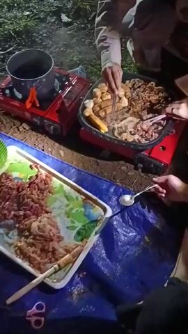 The video depicts a group of people camping and grilling food at night. Several individuals are shown gathered around a small, red portable grill, cooking what appears to be meat, sausages, and other items. One person uses tongs to turn the food on the grill, while another stirs food on a tray. A woman in a hijab is seen crouching near the grill. A fire is burning nearby. The scene is dimly lit, suggesting it is nighttime. The grill and food are placed on a blue tarp laid on the ground.