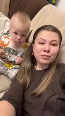 The video shows a woman and a young child sitting on a beige couch. The woman is holding the child on her lap and talking to the camera. The child is wearing pajamas with a dinosaur pattern. The woman is wearing a brown long-sleeved shirt. She has long blonde hair and is wearing gold earrings. Behind them is a pillow with writing on it. The woman holds up one finger and smiles.