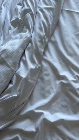 The video opens with a close-up of white, wrinkled bedsheets with a striped pattern. The camera then pans away from the bed to reveal a messy hotel room. Clothes are strewn on a chair and a suitcase is open on the floor. The camera then moves towards a window, through which a Ferris wheel and the ocean are visible. The window is partially open, and a black railing with an ornate design is in the foreground. Cars can be seen driving on a road in front of the Ferris wheel. The sky is clear and blue.