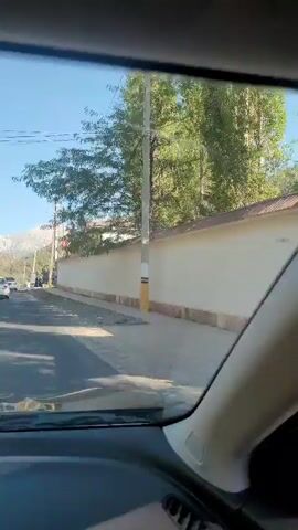 The video shows a point-of-view perspective from inside a vehicle driving on a paved road. The environment appears to be rural or suburban, with a clear blue sky and green trees visible. A white wall with a stone foundation lines the right side of the road for much of the video. The vehicle passes a building with several flags displayed outside, and then continues down the road, which appears to be a two-lane highway. Mountains are visible in the distance. There are other vehicles on the road, including a white car and a dark SUV. There are also some people visible near the building and on the side of the road. The overall impression is of a sunny day drive through a scenic area.