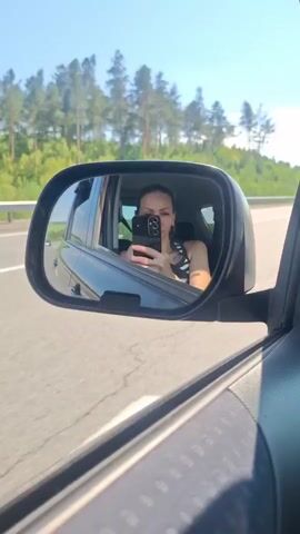A person is seen in the side mirror of a car, holding up a phone and taking a selfie. The car is on a highway, with trees and green hills visible in the background. The sky is blue and clear.