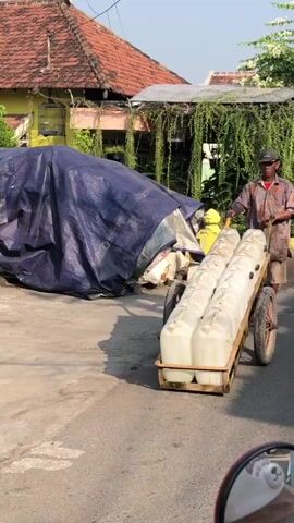 A man pushes a cart filled with large white plastic containers down a street. The cart has two wheels and a wooden frame. The containers are arranged in two rows of three. The man is wearing a cap and a patterned shirt. He is walking on the right side of the street, and there is a blue tarp covering something behind him on the left. The background shows buildings with red tile roofs and green foliage. The lighting suggests it is daytime.