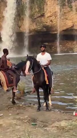 The scene unfolds at the base of a cascading waterfall where a group of people and horses gather on the sandy bank. Several individuals are mounted on horses, engaging in what appears to be recreational riding. Some are dressed in traditional attire while others wear more casual clothing. Individuals on the ground are interacting with the riders and horses, while some are taking photos or videos. The natural backdrop of the waterfall and surrounding vegetation adds to the scenic quality of the gathering. The environment appears to be a popular spot for social and leisure activities.