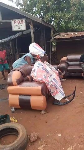 The video shows a man sitting on an orange and brown leather sofa outside. He is wearing a tall, white bag on his head and a robe made from what appears to be Dangote Cement bags. Another man in a blue shirt is standing behind him, appearing to adjust the bag on his head. There are other people around, including a man in a red shirt and blue shorts walking in the background and a person sitting on another brown leather sofa to the right. The setting appears to be outdoors near a building with a sign that reads "P.O.S. AVAILABLE HERE." There is a tire on the ground in the foreground.