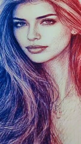 A close-up shot shows a drawing of a woman with her hair divided into blue and red sections. To the right of the drawing, a laptop is open, displaying a reference photo of the same woman. The laptop keyboard is visible at the top of the frame, and the drawing appears to be on a white surface.