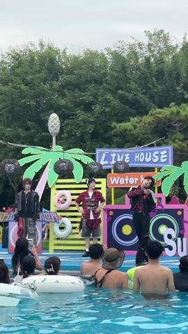 A group of four young men in colorful, stylish clothing are performing on a stage set up at the edge of a swimming pool. The stage is decorated with palm trees, inflatable donuts, and a banner that reads "LIVE HOUSE Water Be". The men are dancing and singing, interacting with each other and the audience, who are mostly women in swimwear, some floating in the pool. The performance appears to be energetic and entertaining. The environment is outdoors, with trees and greenery visible in the background. The overall atmosphere is festive and summery.