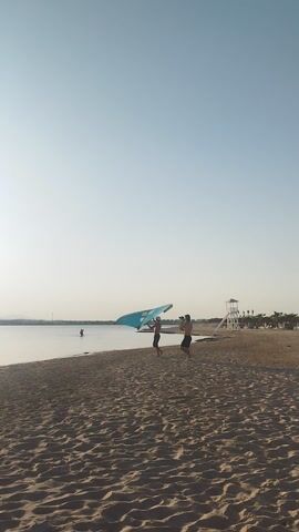 The image captures a wide shot of a sandy beach in Ras Abu Soma, Egypt, during what appears to be either early morning or late afternoon, given the soft, diffused light and clear sky. The horizon line is flat, indicating a calm sea or large bay.

In the mid-ground, two individuals are on the sandy shore next to the water. One person, wearing dark clothing and shorts, is holding a large, brightly colored wing foil, which is predominantly blue with yellow or lime green accents. The wing is partially inflated or spread out, ready for use. The other individual, dressed in dark shorts and a lighter top, stands close by. A board, likely for wingfoiling or paddleboarding, is partially submerged in the shallow water beside them.

In the background, further down the beach, a white lifeguard stand is visible. The sandy beach shows numerous footprints, and the water surface is very calm, reflecting the sky. The overall scene suggests a leisurely water sports activity in a tranquil beach setting. No discernible text is visible in the image.