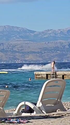The video opens with a view of a beach on a sunny day. In the foreground, two empty white lounge chairs are set up on a pebble beach. Further back, the turquoise water of the sea is visible, with several people swimming. A jet ski is being ridden out on the water, creating a large splash. A person is standing on a concrete pier to the right of the frame. In the background, there is a coastline with buildings and mountains. The camera pans slightly to the left, revealing more of the beach and people relaxing. The jet ski continues to speed across the water, and the person on the pier remains in place. The overall scene depicts a lively beach atmosphere with water sports and relaxation.