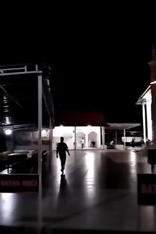 The video begins with a dark shot of a pathway leading to a building. A person walks away from the camera on the left side of the screen. The camera pans to the right, revealing a brightly lit white building with minarets and arched windows. The building appears to be a mosque. A sign in red text reads "BATAS SUCI" which translates to "HOLY BOUNDARY." Two people, possibly a child and an adult, are visible near the front of the building. The scene is illuminated by artificial lighting, creating reflections on the wet ground.