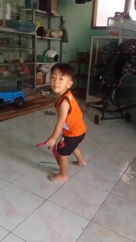 A young child wearing an orange tank top with Elmo on it and black shorts with red trim stands in a room with tiled flooring. The child holds a brown stick-like object, possibly a chocolate bar, and appears to be eating it. A red ribbon or streamer is on the floor to the child's right. In the background, there are glass display cabinets filled with various items, a large black speaker, a blue toy car, and a stroller. The walls are painted green and a window is visible. The child is barefoot and facing forward, looking towards the camera. The child briefly brings the object to their mouth.