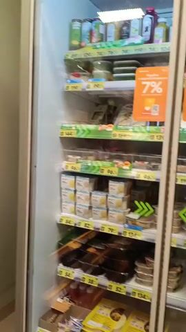 The video pans across a refrigerated display case in a grocery store, likely a convenience store, filled with pre-packaged food items. The items are organized on shelves and include salads, sandwiches, prepared meals, and drinks. Several price tags and promotional signs are visible, including ones indicating a 7% discount and another advertising a sale on ready-made meals up to 40% off. The text on the glass door of the refrigerator appears to be in Russian, with some phrases visible like "Можно поесть" (You can eat) and "с собой" (takeaway). To the right of the display case, shelves of bottled drinks are visible, along with a shopping cart. The overall impression is of a well-stocked refrigerated section in a retail environment.