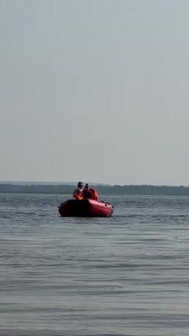 Two people are in a small red inflatable boat on the water. They are both wearing orange life vests. The person in the front is smaller and appears to be a child, while the person in the back is larger and appears to be an adult. The boat is moving slowly through the water. The background shows a hazy horizon line with land and trees. The sky is a pale gray.