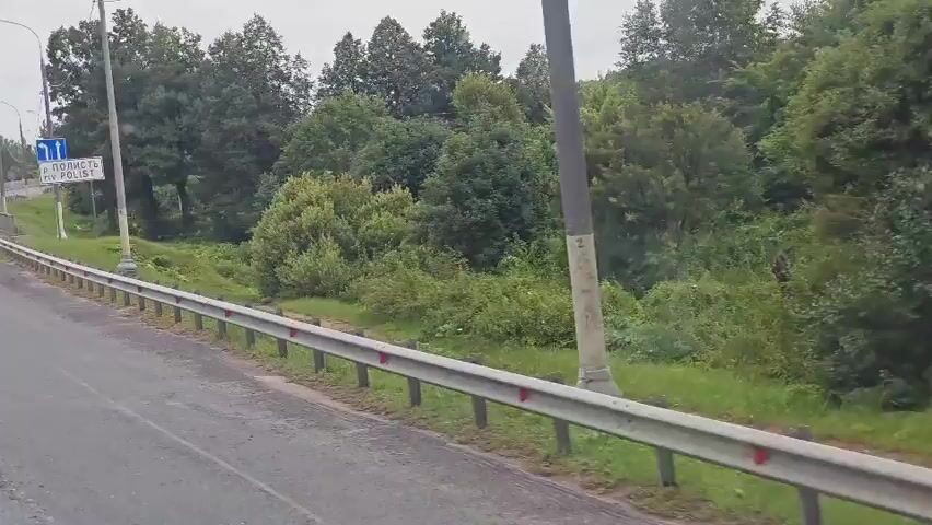 A truck is driving down a road past a guardrail and trees. There are road signs in the background.