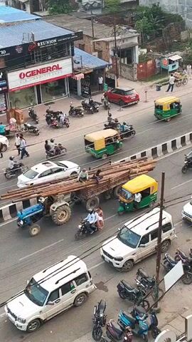 The video shows a busy street scene from an elevated viewpoint. There are multiple vehicles on the road, including cars, motorcycles, and auto-rickshaws. A tractor is pulling a trailer loaded with long wooden poles. People are seen walking on the sidewalk and riding on motorcycles. Buildings line the street, with shops and signs visible. The overall impression is of a typical urban environment with a lot of activity.