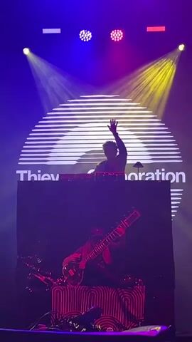 A vertical, medium shot shows a live music performance on a dimly lit stage. In the foreground, a person wearing a hat and sunglasses plays a sitar. Behind them, a DJ stands at a console with equipment, their arm raised. A large, circular backdrop with horizontal lines and the text "Thievery Corporation" is visible behind the DJ. Stage lighting includes yellow and purple spotlights.