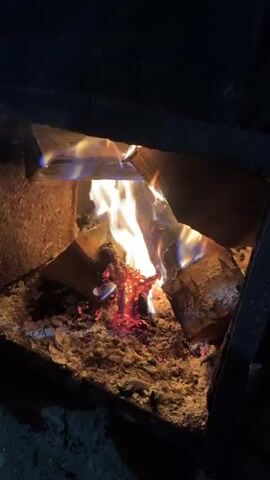The video depicts a fire burning within an enclosed hearth or fireplace. The flames are a combination of yellow, orange, and blue, consuming several pieces of wood. Below the burning wood is a bed of ash and glowing embers. The surrounding walls of the hearth are dark and appear to be made of stone or brick. The video is shot from a stationary position, offering a clear view of the burning fire.