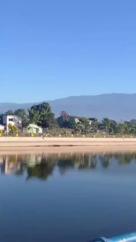 A group of people are walking and jogging on a paved path next to a body of water. In the background, there are trees, buildings, and mountains. The sky is clear and blue. The scene appears to be a park or recreational area. The video pans to the right, following the people as they move along the path. The path is bordered by a railing with blue and yellow accents.