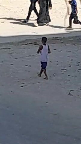 In a dusty, outdoor environment, several people are walking and interacting. In the background, two individuals, one in black attire and another in a dark hijab, walk alongside a boy in a blue shirt carrying a dark bag. To the right, a young boy in a white tank top and blue shorts is standing and appears to be in motion. In the foreground, another boy in a striped shirt and jeans is walking towards the left. Further back and to the right, a person in a dark hijab and grey headscarf is visible, their right arm extended. The scene is set against a backdrop of what appears to be buildings or stalls with signs in an unknown language.
