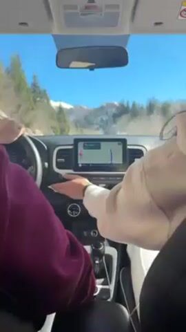A car drives on a paved mountain road between snow-covered slopes. The sky is blue. A person in the driver's seat, wearing a magenta sweatshirt, grips the steering wheel with both hands. A person in the passenger seat, wearing a white hoodie, gestures toward the car's GPS screen. The GPS screen shows a map with a blue route highlighted. The road ahead has patches of snow on the shoulders. A yellow triangular road sign stands on the right shoulder of the road. Trees are visible on the hills. The car is a Hyundai Venue, which can be seen on the GPS screen. The rearview mirror shows a glimpse of the road and the mountain range behind.