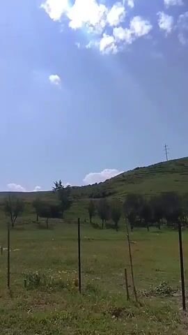 The video captures a bright, sunny day with a clear blue sky and scattered white clouds. The scene is a hilly, grassy landscape with a few scattered trees and a fence. In the distance, a tall, slender tower, possibly a cell tower or transmission tower, stands on top of a hill. The camera pans slightly, showing a small, open-sided structure with a wooden roof on the left side of the frame. The ground in front of the structure appears to be dirt or gravel. The overall impression is one of a rural or semi-rural outdoor setting.