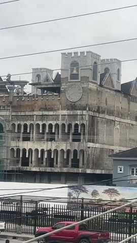 The video shows a large, unfinished building with a castle-like facade, including battlements and arched windows. A large clock face and a bell are visible on the upper portion of the building. The building appears to be under construction, with scaffolding and exposed concrete visible. In the foreground, there is a street with several cars, including a red pickup truck. There are also several street vendors with stalls selling various goods. The sky is overcast. The video appears to be shot from an elevated position, possibly a balcony or another building.