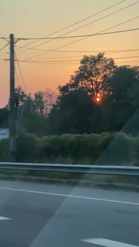 This is a video taken from inside a moving vehicle, looking out the window. The time is either sunrise or sunset, as indicated by the orange and pink hues in the sky. In the foreground, a road and guardrail are visible, suggesting the vehicle is on a highway. Across the road, there is a large white truck trailer with text that appears to read "ACHAT VENTE PALETTE" which translates to "buying selling pallets." Behind the truck, there is a line of trees and some vegetation. Power lines are strung across the sky. There are no people visible in the video. The camera is slightly angled and there are reflections on the window. The overall impression is a fleeting glimpse of a roadside scene during twilight.