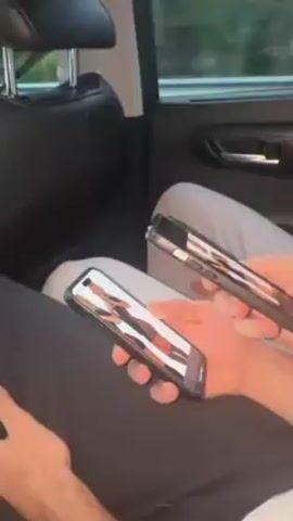 The video shows the interior of a car, presumably the back seat. A person's hands are visible holding a phone and a glass bottle. Another person with curly hair is seen looking at their phone in the front seat. The overall environment suggests a casual car ride.