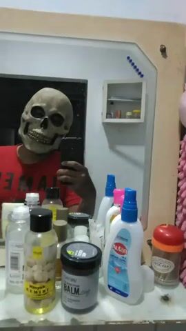 A person wearing a skeleton mask and a red t-shirt with the word "JAMES" visible is taking a selfie in a bathroom mirror. The person is holding a black phone, and their face is obscured by the mask. The bathroom counter is cluttered with various bottles and containers, including Autan lotion and other skincare products. A small, white cabinet with items inside is visible on the wall in the background, as is a mirror with decorative details. The overall lighting is moderate, and the focus is on the masked individual. The camera is handheld and slightly shaky.