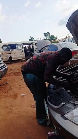 The video captures a male mechanic actively working on the engine of a silver/champagne-colored Honda sedan in an outdoor auto repair setting in Karshi, Nigeria. The car's hood is open, revealing the engine bay, which appears to be in a state of repair or inspection.

The man, with a dark complexion, is wearing a short-sleeved, patterned shirt (red, black, and green floral/camouflage-like design) and teal-colored pants. He is bent over the front of the car, focusing intently on the engine components, with both hands inside the engine bay. A car battery, predominantly red and black, is placed on the dirt ground directly in front of the car, seemingly connected to the vehicle. Visible text on the battery includes "RUI POWER" and "Maintenance Free," along with some partially visible Chinese characters.

The scene is set on a dirt ground under bright daylight, indicating daytime. In the background, several other vehicles are parked, including a white van and another silver sedan with its hood also open, further reinforcing the auto workshop environment. Trees and some buildings are visible in the distance. The Honda car displays a "HONDA" logo on its grille and a green Nigerian license plate, though the specific characters are not fully legible. The man's shoe features a "NIKE" swoosh. The overall atmosphere suggests a busy, functional repair site.