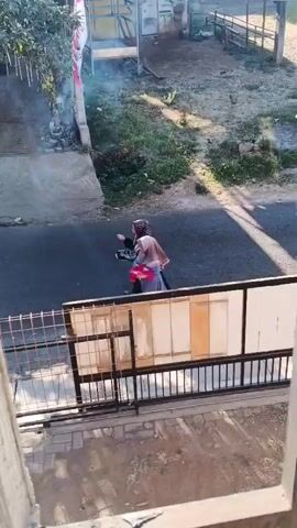 At timestamp 00:00, a person is walking on the asphalt road, carrying a red bag and a tray with some items on it. A flagpole with a flag is visible on the left side of the road. At timestamp 00:01, a motorcycle with a rider wearing a yellow shirt and a helmet is passing by on the same road. At timestamp 00:02, the motorcycle has passed, and the road is empty except for the person from the beginning of the video who is still walking. At timestamp 00:03, the person has moved further down the road, and a motorcycle with two riders is approaching. The rider in front is wearing a blue shirt, and the passenger is wearing a pink headscarf. At timestamp 00:04, the motorcycle with two riders is passing the person walking on the road. The scene appears to be an outdoor environment with some buildings and vegetation in the background, possibly in a residential area. The lighting suggests it is daytime.